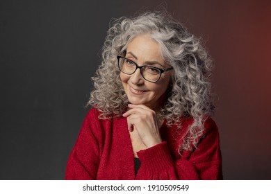 Studio Portrait Of A Smiling Elderly Woman 60-65 Years Old In A Red Sweater And Glasses, Gray Curly Long Hair, On A Colored Background. Concept: Stylish Pensioners Of Model Appearance, Active Life.