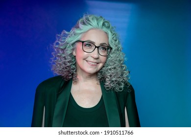 Studio Portrait Of A Smiling Elderly Woman Of 60-65 Years Old In A Strict Black Suit And Glasses, With Gray Curly Long Hair, On A Colored Background. 