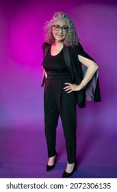 Studio Portrait Of A Smiling Elderly Woman 60-65 Years Old With Glasses, With Gray Curly Long Hair, On A Colored Background, Concept: Stylish Pensioners And Grandmothers Of Model Appearance, Active.