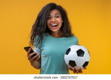 Studio portrait of  smiling lucky football fan girl holding ball in her hand and making bets on favorite team at bookmaker's website using smartphone. Isolated on yellow background.