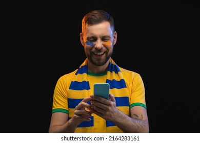 Studio portrait of smiling lucky football fan making bets on favorite team at bookmaker's website using smartphone, isolated over black background. Sport betting, gambling, money win concept. - Shutterstock ID 2164283161