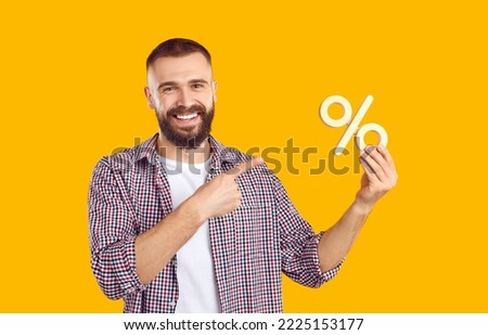 Studio portrait of smiling man with percentage sign. Happy young man in casual shirt isolated on orange yellow background advertising sale and pointing at discount percent symbol in his hand
