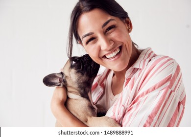 Studio Portrait Of Smiling Young Woman Holding Affectionate Pet French Bulldog Puppy