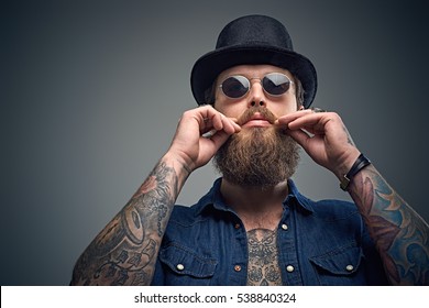 Studio portrait of stylish bearded male with a tattoo on his chest, dressed in a denim shirt, cylinder hat and sunglasses isolated on grey background.