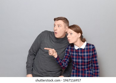 Studio portrait of surprised couple, with widened eyes and open mouths, woman is pointing with index finger at something extraordinary, both are standing together over gray background
