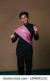 Studio Portrait Of A Tall And Handsome Malay Asian Man In A Traditional Black Baju Melayu Outfit With A Festive Pink Sash Over His Shoulder. He Is Smiling, Laughing And Dancing In Celebration. 