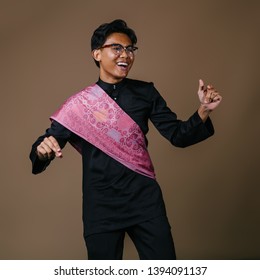 Studio Portrait Of A Tall And Handsome Malay Asian Man In A Traditional Black Baju Melayu Outfit With A Festive Pink Sash Over His Shoulder. He Is Smiling, Laughing And Dancing In Celebration. 