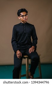 Studio Portrait Of A Tall And Handsome Malay Asian Man In A Traditional Black Baju Melayu Outfit Sitting On A Chair. He Has A Neutral Expression And Looks Intelligent And Educated With His Spectacles.