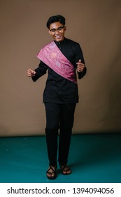 Studio Portrait Of A Tall And Handsome Middle Eastern Man In A Traditional Black Baju Melayu Outfit With A Festive Pink Sash Over His Shoulder. He Is Smiling.