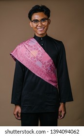 Studio Portrait Of A Tall And Handsome Middle Eastern Man In A Traditional Black Baju Melayu Outfit With A Festive Pink Sash Over His Shoulder. He Is Smiling.