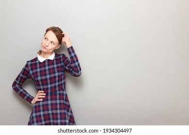 Studio portrait of thoughtful puzzled blond girl wearing checkered dress, scratching her head with finger, being deep in thoughts, trying to solve issue, standing over gray background, with copy space - Powered by Shutterstock