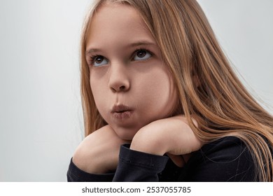 Studio portrait of a thoughtful young girl with long blond hair, resting her chin on her hands, her puffed cheeks and contemplative gaze suggesting deep thought or boredom - Powered by Shutterstock