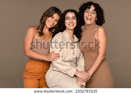 Studio portrait of three beautiful women in their 20s and 30s posed smiling and laughing on a neutral background
