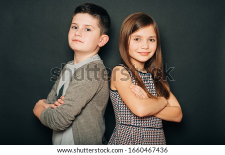 Studio portrait of two cute kids, standing back to back, arms crossed, posing on black background