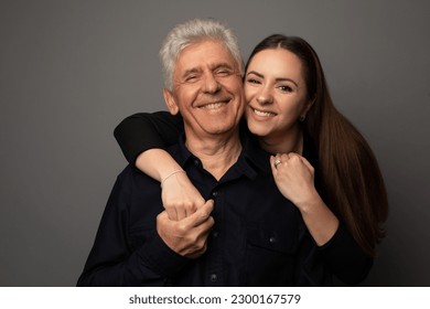 Studio portrait of a Ukrianian father in his 60s and his adult daughter in her late 20s. They are both wearing black and the daughter is hugging her father. The background is grey. - Powered by Shutterstock