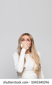 Studio portrait of unhealthy cute blonde female in white top with napkin blowing nose, looks up to the source of the allergy, source of unpleasant odor/Place for advertising/Rhinitis, allergy concept - Shutterstock ID 1045340296