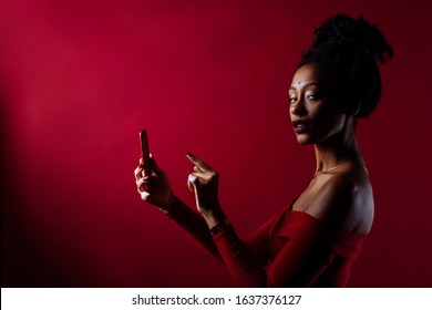 Studio portrait of a woman with dreadlocks wearing a sparkling dress in a studio portrait with red background. Texting on her mobile phone