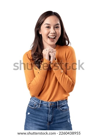 A studio portrait of a young Asian Indonesian woman wearing an orange long-sleeve shirt looks happy as she smiles while her hands are clasped in front of her chest. Isolated on a white background.