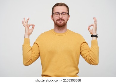 Studio Portrait Of Young Bearded Male Student Wears Yellow Sweater Keeps Calm, Keeps Eyes Closed And Showing Yoga Mudra Sign, Isolated Over White Background