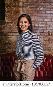 Studio Portrait Of A Young, Beautiful, Elegant And Intelligent Looking Indian Asian Woman In A Striped Business Shirt And Casual Khakis Smiling As She Poses For Her Headshot Indoors.