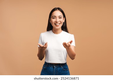 Studio portrait of a young, beautiful woman with long dark hair, wearing a white t-shirt and jeans, smiling and gesturing with her hands outstretched against a beige background - Powered by Shutterstock