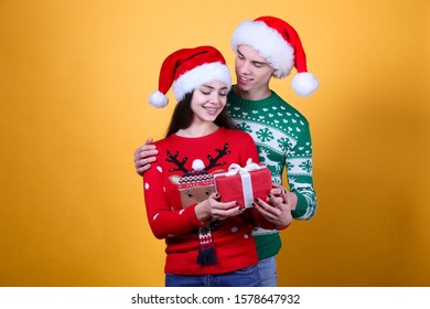 Studio Portrait Of Young Couple, Boyfriend & Girlfriend Wearing Santa Claus Hat & Ugly Christmas Sweater. Holiday Outfit W/ Snowflake Pattern Print. Close Up, Copy Space For Text, Isolated Background.