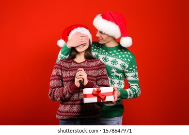 Studio Portrait Of Young Couple, Boyfriend & Girlfriend Wearing Santa Claus Hat & Ugly Christmas Sweater. Holiday Outfit W/ Snowflake Pattern Print. Close Up, Copy Space For Text, Isolated Background.