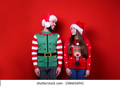 Studio Portrait Of Young Couple, Boyfriend & Girlfriend Wearing Santa Claus Hat & Ugly Christmas Sweater. Holiday Costume, Elf Vest Print. Close Up, Copy Space For Text, Isolated Background.