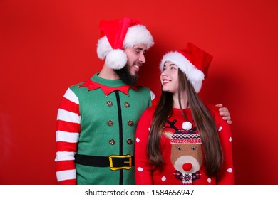 Studio Portrait Of Young Couple, Boyfriend & Girlfriend Wearing Santa Claus Hat & Ugly Christmas Sweater. Holiday Costume, Elf Vest Print. Close Up, Copy Space For Text, Isolated Background.