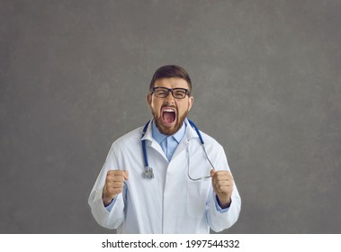 Studio Portrait Of Young Doctor Stressed By Problem At Work Going Postal And Shouting. Angry Frustrated Mad Crazy Furious General Practitioner Clenching Fists And Screaming Isolated On Gray Background