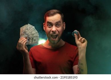 Studio Portrait Of Young Euphoric Man Posing With A Stack Of Poker Chips And Ward Of Dollar Banknotes In Hands Over Black Background With Smoke Effect. Gambling, Sport Betting, Poker, Casino Concept.