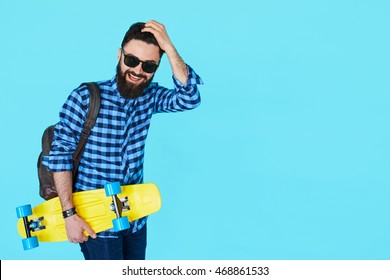 Studio Portrait Of Young Fashionable Hipster Man Posing Isolated Over Blue Background With Copy Space. Bearded Male Holding Yellow Skateboard And Smiling