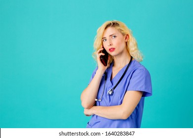 Studio Portrait Of A Young Female Doctor Or Nurse Wearing Blue Scrub Uniform And Stethoscope Talking On Mobile Phone With Concerned Expression