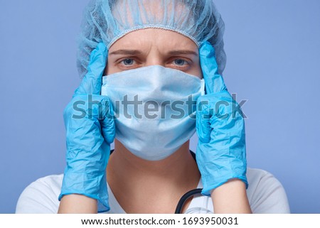 Studio portrait of young girl having headache, holding hands on temples, wearing medical flu mask, rubber gloves and disposable hat, posing isolated over blue background. Healrh care concept. Stock photo © 