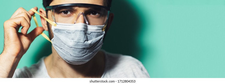 Studio Portrait Of Young Guy Putting On Medical Face Mask, Wearing Safety Goggles Against Coronavirus On Background Of Aqua Menthe Color With Copy Paste.