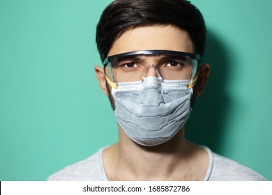 Studio Portrait Of Young Guy Wearing Medical Flu Mask And Safety Goggles, Coronavirus Prevention, Isolated On Background Of Cyan, Aqua Menthe Color.