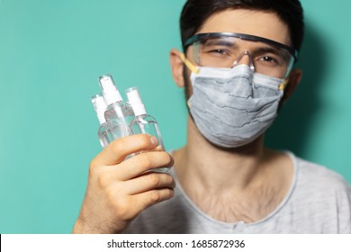 Studio Portrait Of Young Guy Wearing Medical Flu Mask And Safety Goggles Against Coronavirus, Holding Dispenser Bottles With Sanitizer Antiseptic Gel, On Background Of Cyan, Aqua Menthe Color.