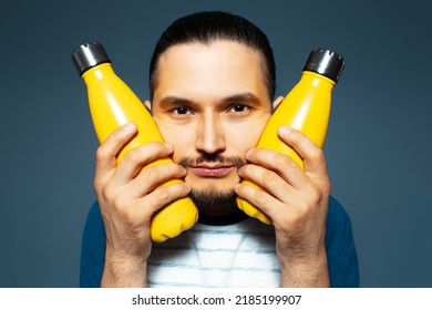 Studio Portrait Of Young Handsome Man, Holding Two Yellow Eco Bottles Close To His Face.