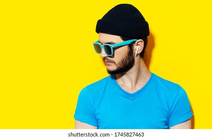 Studio Portrait Of Young Hipster Guy Using Wireless Earphones, Wearing Blue Shirt, Cyan Sunglasses And Black Beanie Hat On Background Of Yellow Color With Copy Space.