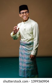 Studio Portrait Of A Young Malay Asian Man In A Baju Melayu, Sampin And Songkok Hat. He Is Smiling Happily And Confidently As He Poses For His Head Shot In A Studio. 