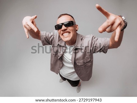 Studio portrait of a young man in gray shirt and sunglasses making expressive rapper hand gestures and facial expressions. Fisheye top-angle lens creates strong body distortion on gray background.