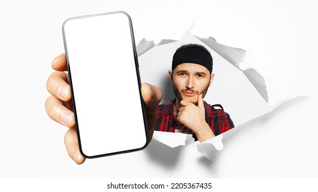 Studio Portrait Of Young Man, Showing Big Smartphone With Blank On Screen Through Torn Paper Hole. White Background. Holding Hand On Chin, Wearing Black Band On Head.