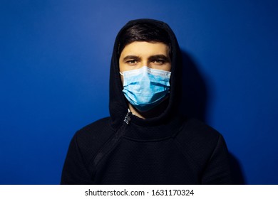 Studio Portrait Of Young Man Wearing Medical Flu Mask And Hoodie Sweater. Background Of Phantom Blue Color.