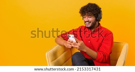 Studio portrait of young middle eastern guy in wireless headphones isolated on orange wall, looking at mobile phone, enjoying listening to favorite music soundtrack