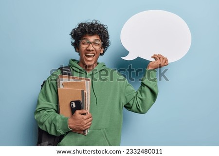 Studio portrait of young smiling broadly glad Hindu male student standing in centre with books and smartphone at hand holding big white speech bubble for your promotion isolated on blue background