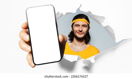 Studio Portrait Of Young Smiling Sportsman, Showing Big Smartphone With Blank On Screen Through Torn Paper Hole. White Background. Wearing Yellow Band On Head And Shirt.