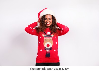 Studio Portrait Of Young Woman With Dark Skin And Long Curly Hair Wearing Tight Santa Claus Hat And Christmas Outfit. Ugly Sweater Concept. Close Up, Copy Space For Text, Isolated Background.