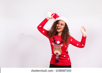 Studio Portrait Of Young Woman With Dark Skin And Long Curly Hair Wearing Tight Santa Claus Hat And Christmas Outfit. Ugly Sweater Concept. Close Up, Copy Space For Text, Isolated Background.