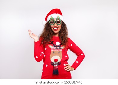 Studio Portrait Of Young Woman With Dark Skin And Long Curly Hair Wearing Tight Santa Claus Hat And Christmas Outfit. Ugly Sweater Concept. Close Up, Copy Space For Text, Isolated Background.