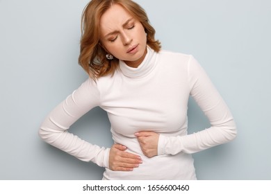 Studio portrait of young woman having a stomachache, menstruation pain or cramps, isolated on grey background. Chronic gastritis, period menstruation, female health problem, aching belly, gynecology